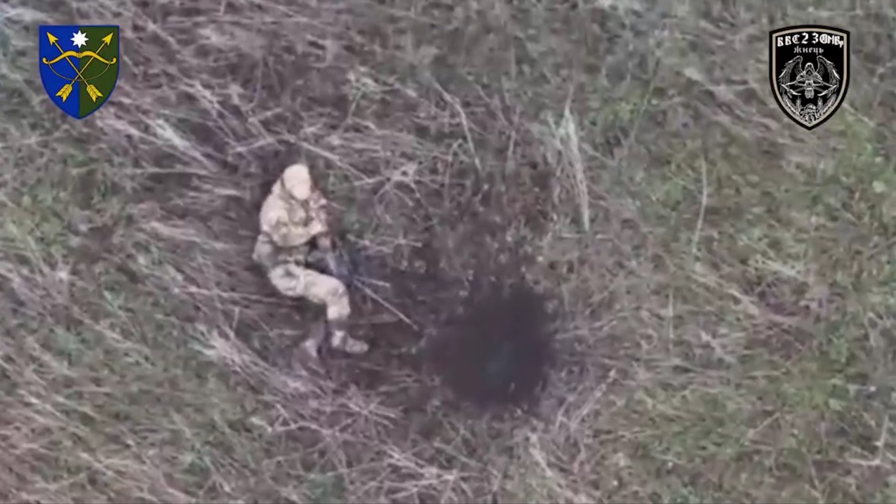 Russian is lying on the ground in the middle of open field, apparently injured next to his comrade who looks dead. Drone dropped grenade lands really close, he rolls over to dodge it narrowly, grabs his AK and shoots himself blowing away his hat