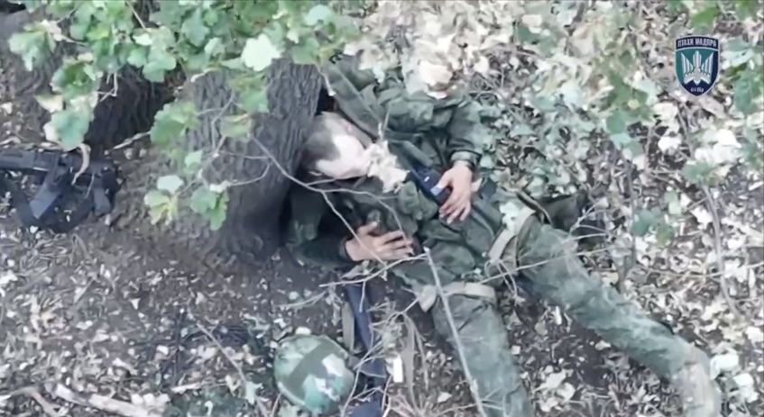 Russian soldier walks along treeline, video fast forwards to him sitting under the tree under drone drop attack. Camera zooms in as he calmly takes grenade, puts it under his vest and detonates. Later on camera pans over gruesome aftermath