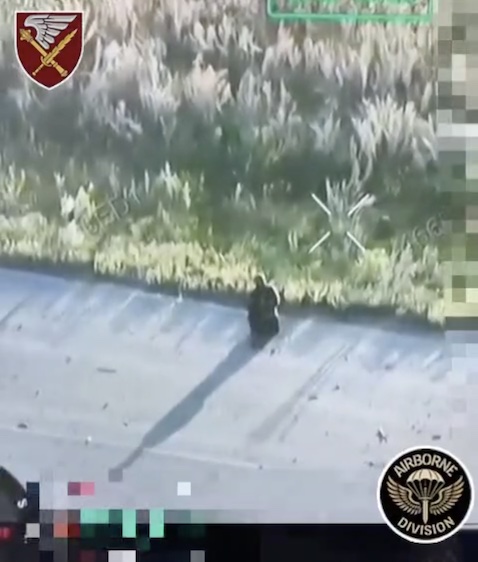Short clip featuring Russian soldier standing kneeled on the paved road, pointing his rifle under his chin and pulling the trigger