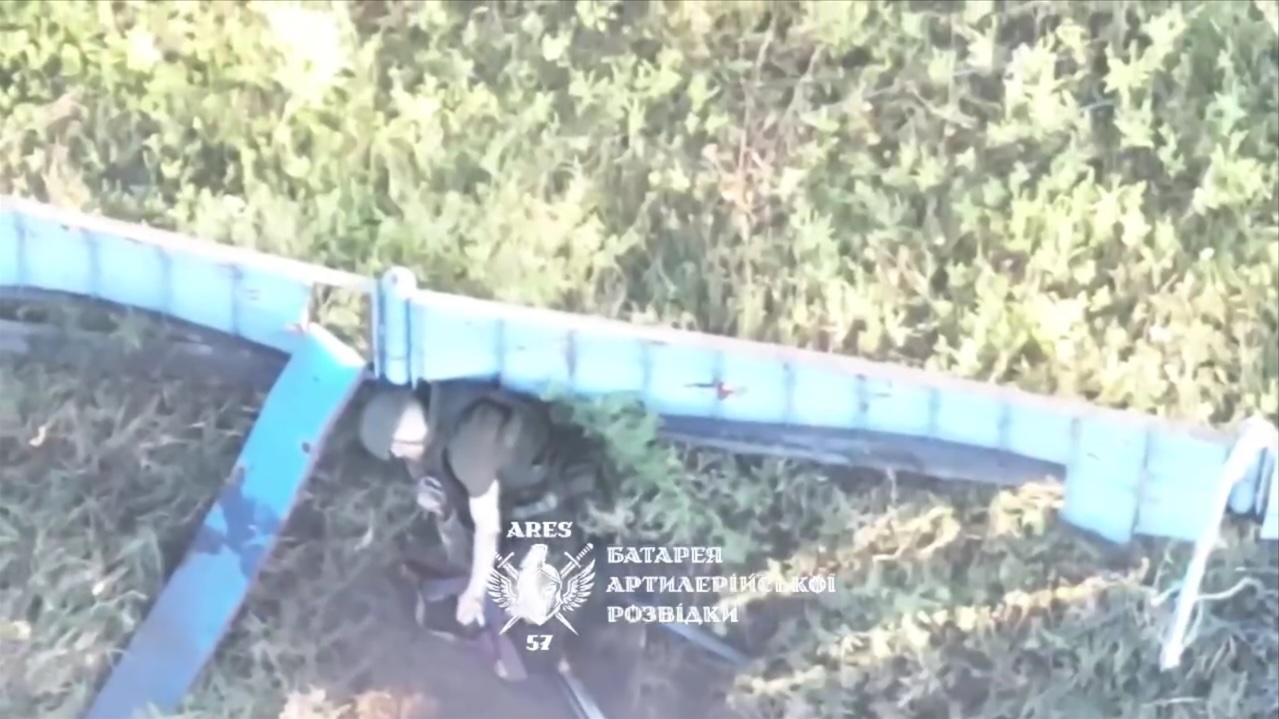 Russian is lying besides what looks like metal fence. After receiving drone drop, he tries to reach out for something in his hip belt, eventually grabs AK and shoots himself