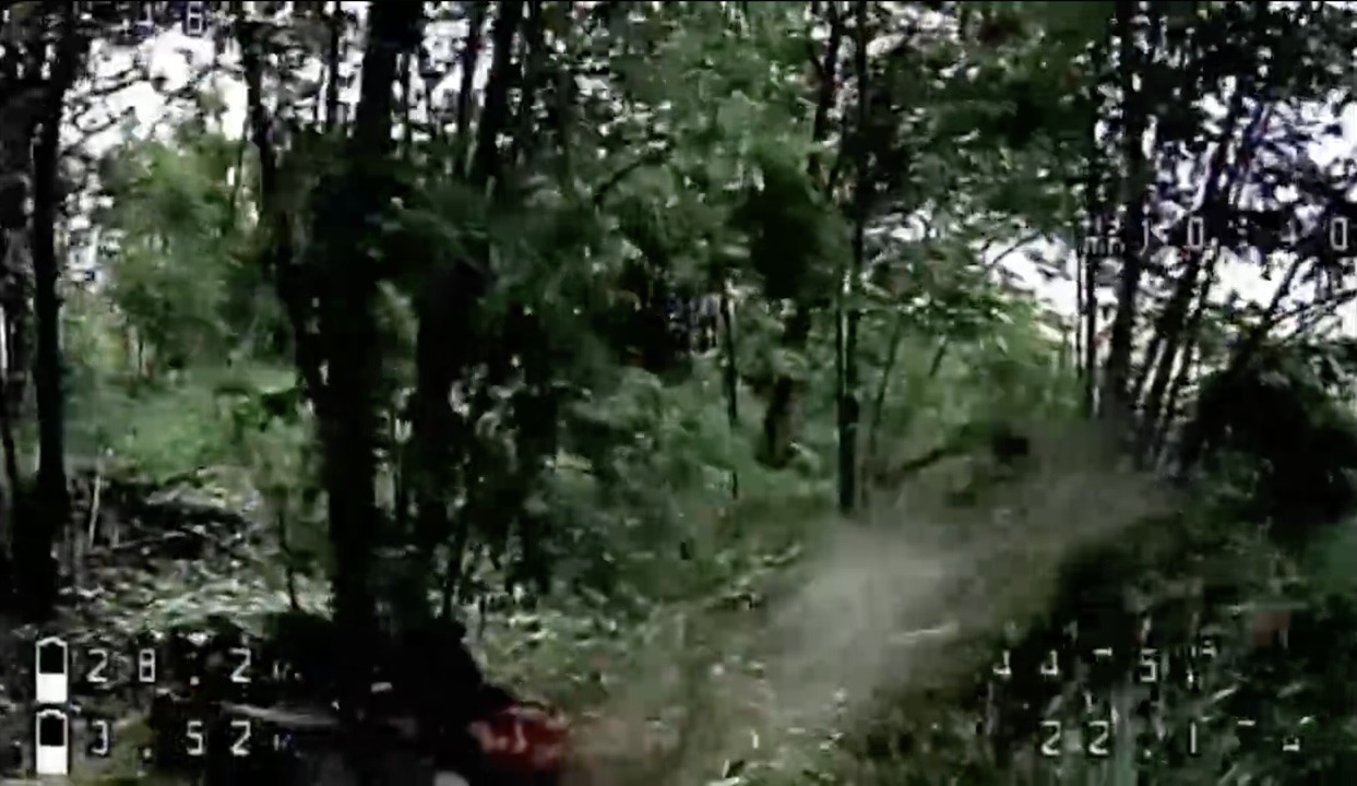 FPV seeks enemy in a tree line with a lot of foliage. Just as it approaches Russian soldier lying on his side under the tree, he shoots himself wit hhis AK on full auto